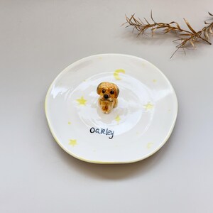 Peut inclure: Une assiette en c&eacute;ramique blanche avec un bord jaune, d&eacute;cor&eacute;e d'&eacute;toiles et de croissants de lune jaunes. Une petite figurine de chien d&eacute;taill&eacute;e est au centre. Le nom "oakley" est &eacute;crit en script noir sur l'assiette.