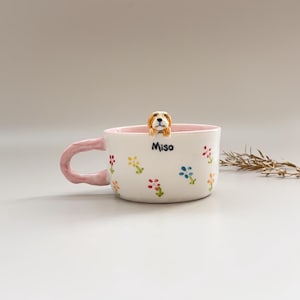 Puede incluir: Taza de cer&aacute;mica blanca con interior y asa rosas, adornada con dise&ntilde;os florales coloridos. Una peque&ntilde;a y detallada figura de perro asoma por el borde. La taza tiene el nombre "Miso".