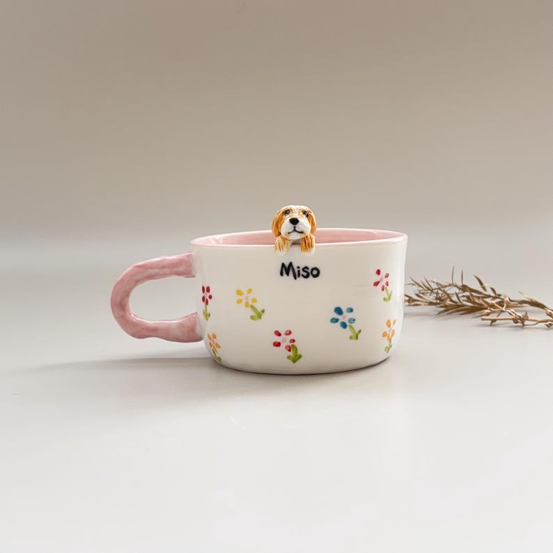 Puede incluir: Taza de cer&aacute;mica blanca con interior y asa rosas, adornada con dise&ntilde;os florales coloridos. Una peque&ntilde;a y detallada figura de perro asoma por el borde. La taza tiene el nombre "Miso".