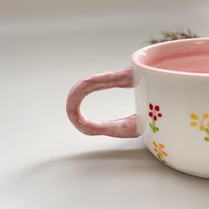 Puede incluir: Taza de cer&aacute;mica blanca con interior y asa rosas. La taza est&aacute; decorada con peque&ntilde;as flores de colores. El asa tiene un dise&ntilde;o curvo y grueso. La taza probablemente se utiliza para beber bebidas.