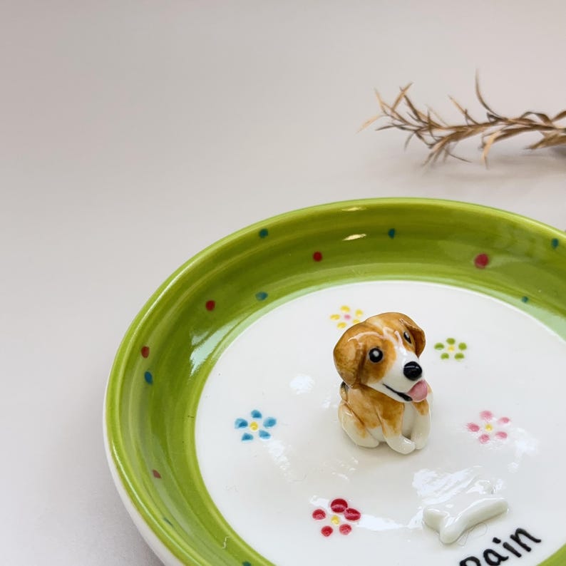 May include: A ceramic dog figurine with brown and white fur sits on a white plate with a green rim. The plate is decorated with colorful polka dots and floral designs. A small bone-shaped object is also on the plate.