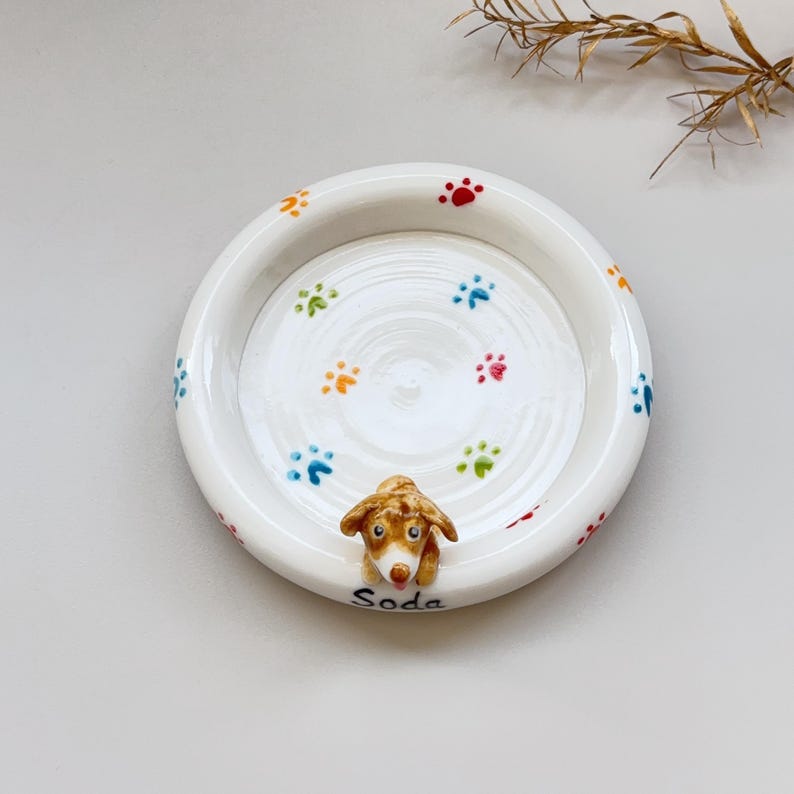 K&ouml;nnte beinhalten: Eine wei&szlig;e Keramikschale mit bunten Pfotenabdr&uuml;cken und einer kleinen, braun-wei&szlig;en Hundefigur. Der Hund streckt die Zunge heraus. Das Wort "Soda" ist auf der Schale geschrieben.