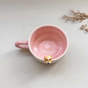 Puede incluir: Una taza de cer&aacute;mica rosa con un detalle de cara de perro en el borde. La taza tiene un asa redondeada y un interior rosa claro con un dise&ntilde;o en espiral. La cara del perro es marr&oacute;n y blanca con ojos y nariz negros.
