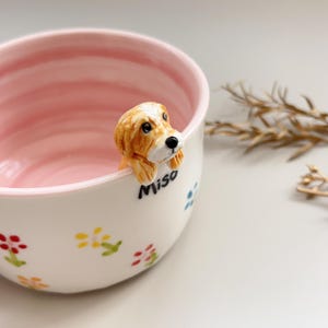 Puede incluir: Un cuenco de cer&aacute;mica blanco con interior rosa y una figura decorativa de perro tridimensional en el borde. El cuenco est&aacute; adornado con coloridos motivos florales y el nombre "Miso" est&aacute; escrito en el lateral.
