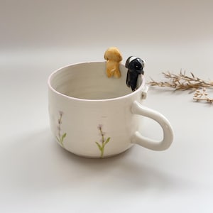 May include: White ceramic mug with a light pink rim and a curved handle. The mug is decorated with lavender sprigs and has two small dog figurines perched on the rim. One dog is tan, the other is black and white.