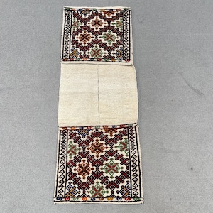 May include: An off-white textile with geometric patterns in red, brown, green, and blue. The top and bottom sections have detailed designs, while the central area is plain. The edges are bordered with a black and white pattern. The textile is rectangular.