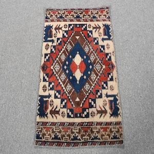 May include: A rectangular area rug featuring a central diamond motif in deep blue and brown, surrounded by geometric patterns in red, cream, and dark blue. The border displays intricate repeating shapes in brown, red, and blue on a cream background.