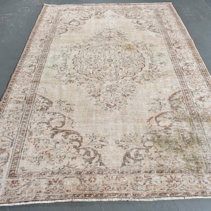 May include: A rectangular area rug featuring a central medallion and intricate floral patterns in shades of beige, brown, and muted green. The rug has a wide decorative border with repeating motifs and a weathered, distressed texture throughout.