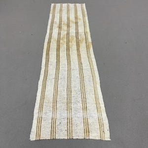 May include: A long, rectangular runner rug featuring a textured, off-white base with vertical, parallel stripes in a muted tan or light brown color. The fabric has a visible, coarse weave and frayed edges at the top and bottom ends of the piece.