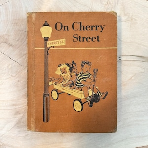 Könnte beinhalten: Ein altes Kinderbuch mit dem Titel "On Cherry Street" mit braunem Einband und einer Illustration von zwei Kindern und einem Hund, die in einem Wagen fahren. Der Wagen wird von einem Stock mit einer Schaufel am Ende gezogen. Die Kinder tragen Freizeitkleidung und der Hund ist braun und weiß.