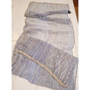 May include: A rectangular light blue scarf featuring a semi-sheer fabric with a repeating pattern of silver-toned swirls and geometric lines. The edges are finished with a delicate cream-colored crocheted lace trim along the length of the scarf.
