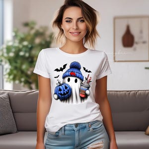 May include: White t-shirt featuring a cartoon ghost wearing a blue knit cap with a red heart, holding a blue pumpkin and a coffee drink. Black bats are flying around the ghost. The shirt is a Halloween-themed design.