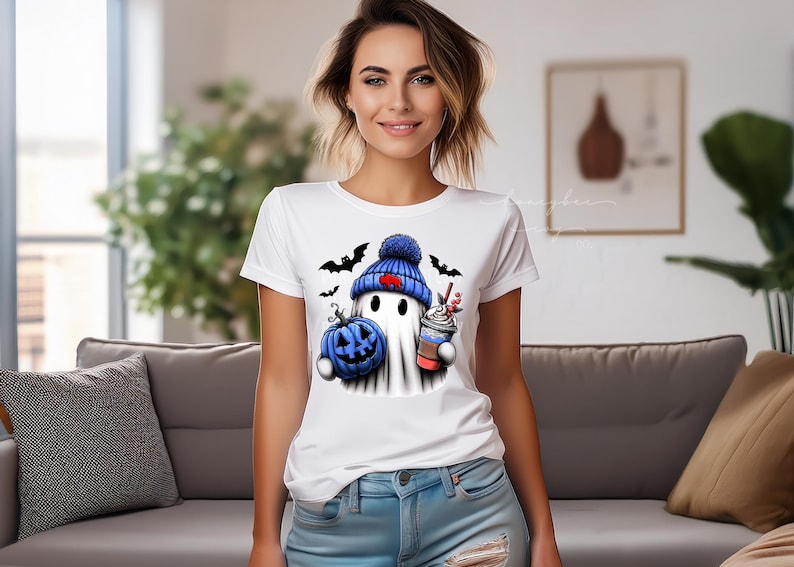 May include: White t-shirt featuring a cartoon ghost wearing a blue knit cap with a red heart, holding a blue pumpkin and a coffee drink. Black bats are flying around the ghost. The shirt is a Halloween-themed design.
