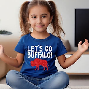 May include: A young person wearing a blue t-shirt with the text "LET'S GO BUFFALO!" in white and a red buffalo graphic. The person is sitting with their legs crossed, wearing blue jeans.
