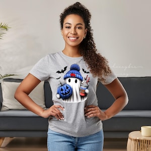 May include: A light gray t-shirt featuring a Halloween-themed graphic. The graphic includes a ghost wearing a blue beanie, holding a blue pumpkin and a coffee cup. Black bats are also included in the design.