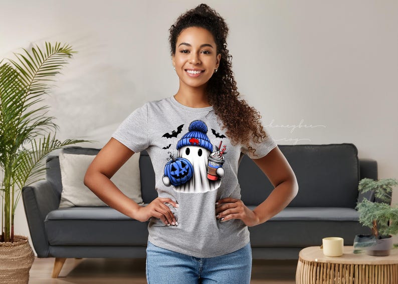 May include: A light gray t-shirt featuring a Halloween-themed graphic. The graphic includes a ghost wearing a blue beanie, holding a blue pumpkin and a coffee cup. Black bats are also included in the design.
