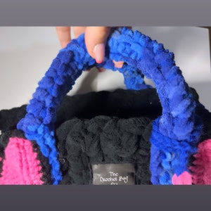 May include: A black crocheted bag with blue and pink accents. The bag has a fluffy texture and a handle. The bag has a tag that says "The Crochet Bag".