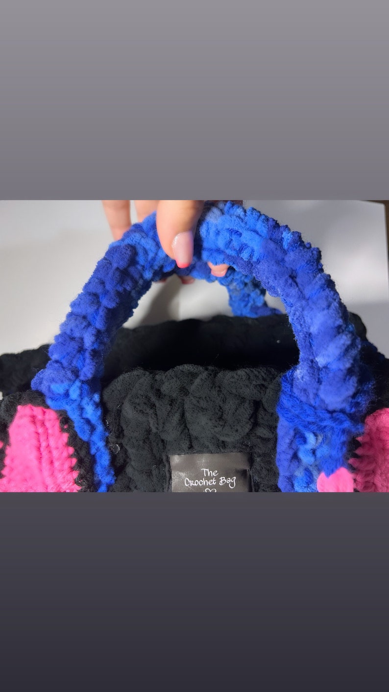 May include: A black crocheted bag with blue and pink accents. The bag has a fluffy texture and a handle. The bag has a tag that says "The Crochet Bag".