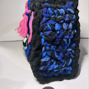 May include: A blue, black, and pink crocheted tote bag with a fringe detail. The bag is made with a thick, textured yarn.