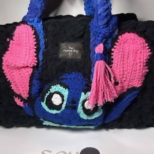 May include: A blue and pink crocheted bag with a cartoon character design. The bag has a fluffy texture and a pink tassel. The bag has a tag that says "The Crochet Bag".