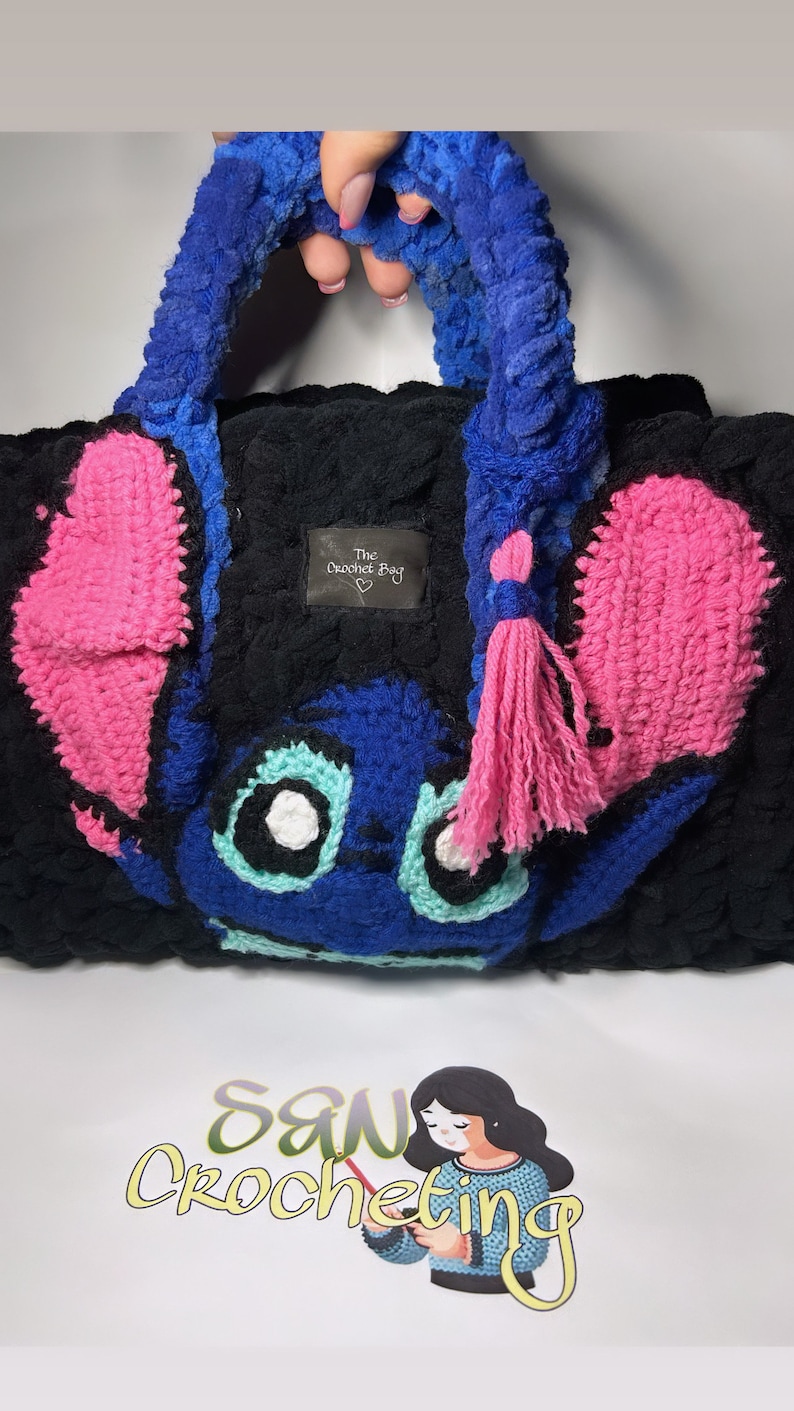 May include: A blue and pink crocheted bag with a cartoon character design. The bag has a fluffy texture and a pink tassel. The bag has a tag that says "The Crochet Bag".