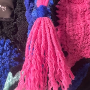May include: A close-up of a pink and blue crocheted bag with a long pink tassel. The bag is made of different colored yarns and has a unique, textured design.