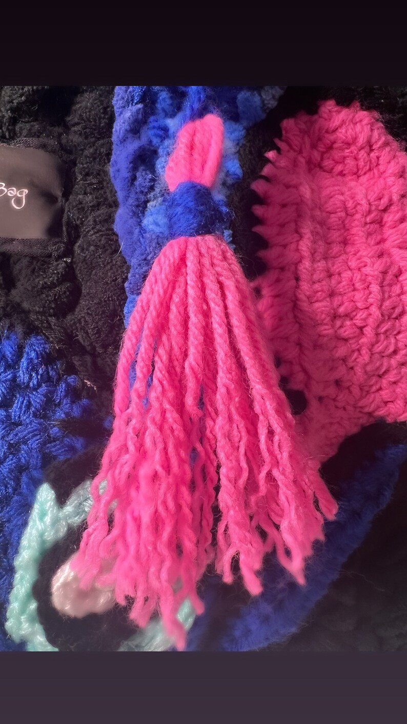 May include: A close-up of a pink and blue crocheted bag with a long pink tassel. The bag is made of different colored yarns and has a unique, textured design.