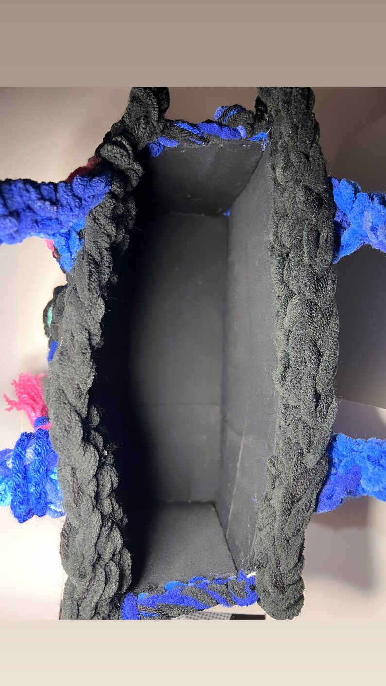 May include: A black crocheted tote bag with blue and pink yarn accents. The bag has a black fabric lining.