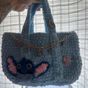 K&ouml;nnte beinhalten: Eine hellblaue, handgefertigte H&auml;keltasche. Die Tasche hat eine goldene Kette und Stitch aus Lilo & Stitch auf der Vorderseite appliziert. Zus&auml;tzliche Charms und Verzierungen sind an der Kette und der Tasche befestigt.