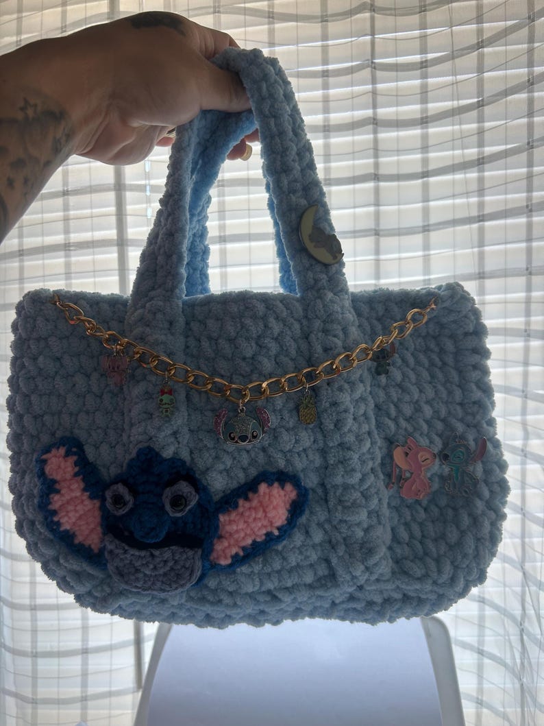 K&ouml;nnte beinhalten: Eine hellblaue, handgefertigte H&auml;keltasche. Die Tasche hat eine goldene Kette und Stitch aus Lilo & Stitch auf der Vorderseite appliziert. Zus&auml;tzliche Charms und Verzierungen sind an der Kette und der Tasche befestigt.