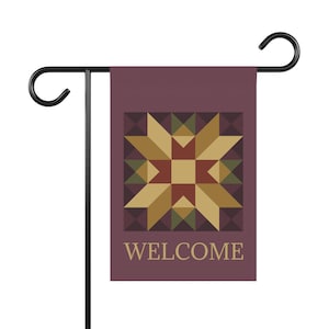 Puede incluir: Una bandera de jardín rectangular de color ciruela intenso con un patrón de estrella geométrico en tonos oro, rojo oscuro y verde oliva. La palabra WELCOME está impresa en una fuente serif de color tostado claro en la parte inferior, colgada de un soporte de metal negro.