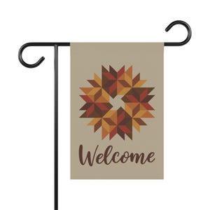 May include: A rectangular garden flag hanging on a black metal stand. The flag is beige and features a central geometric star pattern in shades of brown, orange, and red. Below the star, the word Welcome is printed in a dark brown script font.
