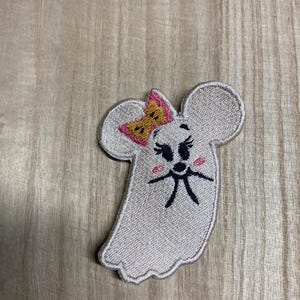 May include: A light gray embroidered patch in the shape of a ghost with a mouse-like face. The ghost has a pink bow, black eyes, and a black mouth. The patch is on a wooden surface.