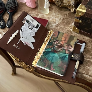 以下が含まれることがあります： 金色のリングが付いた茶色の革製プランナー、ペン、バレリーナのプリント。 プランナーには「N.S.S.」のイニシャルと白いレースの蝶が付いています。 背景には香水瓶とヘアブラシがあります。