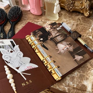 May include: A brown leather planner with gold rings, featuring a print of ballet dancers, a white lace butterfly, a pearl hair clip, and the text N.S.S. LOUIS VUITTON PARIS MADE IN FRANCE. A pen with a brown monogram pen loop is attached.
