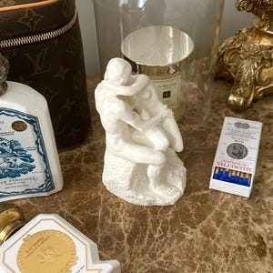 Peut inclure: Sculpture en porcelaine blanche d'un couple qui s'embrasse, sous un d&ocirc;me en verre, sur une surface brune avec un flacon de parfum et une bo&icirc;te d'allumettes.
