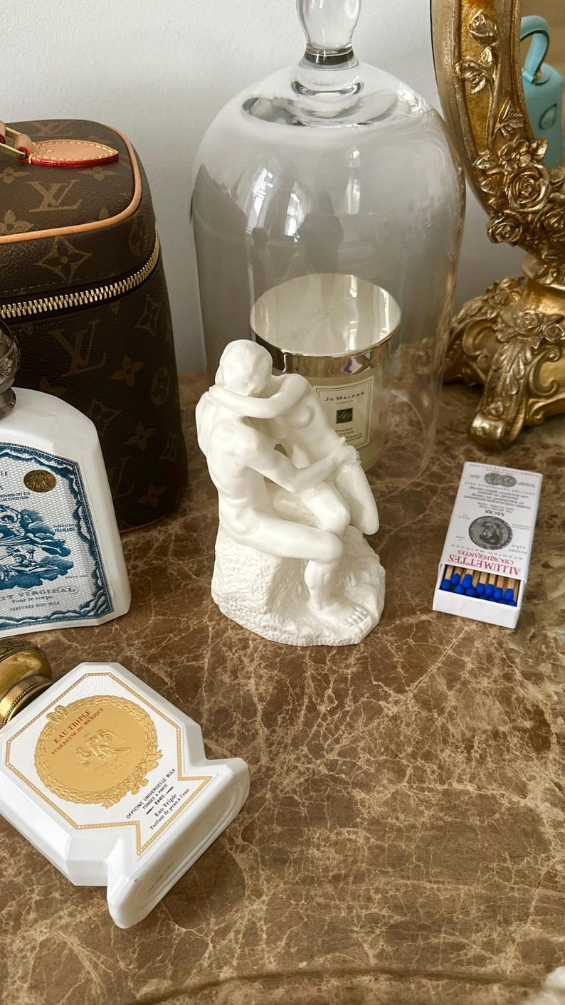 Peut inclure: Sculpture en porcelaine blanche d'un couple qui s'embrasse, sous un d&ocirc;me en verre, sur une surface brune avec un flacon de parfum et une bo&icirc;te d'allumettes.