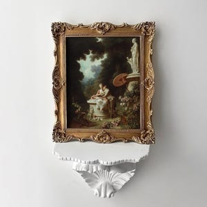 May include: A framed painting with a gold ornate frame depicting a romantic garden scene. The artwork is mounted on a white decorative shelf. The painting features figures, a statue, and greenery, creating a classical aesthetic.