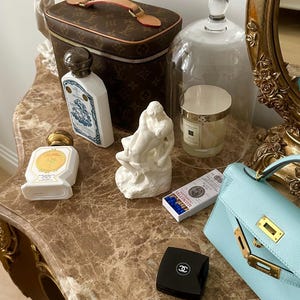 Peut inclure: Une table de toilette avec un design orn&eacute; brun et or. Sur la table se trouvent un n&eacute;cessaire de toilette Louis Vuitton brun et or, un flacon en porcelaine blanche avec un motif bleu et or, un flacon en porcelaine blanche avec un bouchon dor&eacute;, une sculpture en porcelaine blanche de deux personnages, une cloche en verre avec une bougie blanche &agrave; l'int&eacute;rieur, une bo&icirc;te d'allumettes, un compact noir avec un logo Chanel et un sac &agrave; main bleu clair avec des garnitures dor&eacute;es.