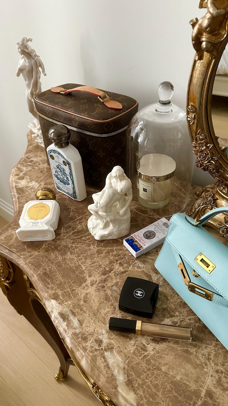 Peut inclure: Une table de toilette avec un design orn&eacute; brun et or. Sur la table se trouvent un n&eacute;cessaire de toilette Louis Vuitton brun et or, un flacon en porcelaine blanche avec un motif bleu et or, un flacon en porcelaine blanche avec un bouchon dor&eacute;, une sculpture en porcelaine blanche de deux personnages, une cloche en verre avec une bougie blanche &agrave; l'int&eacute;rieur, une bo&icirc;te d'allumettes, un compact noir avec un logo Chanel et un sac &agrave; main bleu clair avec des garnitures dor&eacute;es.