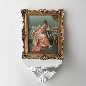 Puede incluir: Una impresión enmarcada de una mujer con un vestido rosa sentada a una mesa, con un marco dorado ornamentado. La impresión está montada en un estante decorativo blanco con un borde festoneado y un diseño de hoja.