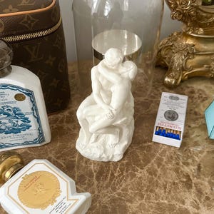 Peut inclure: Une sculpture en porcelaine blanche repr&eacute;sentant un couple en train de s'embrasser, expos&eacute;e sous un d&ocirc;me en verre sur une surface en marbre. La sculpture est entour&eacute;e d'autres objets d&eacute;coratifs, dont un flacon de parfum, une bo&icirc;te d'allumettes et un sac &agrave; main Louis Vuitton.