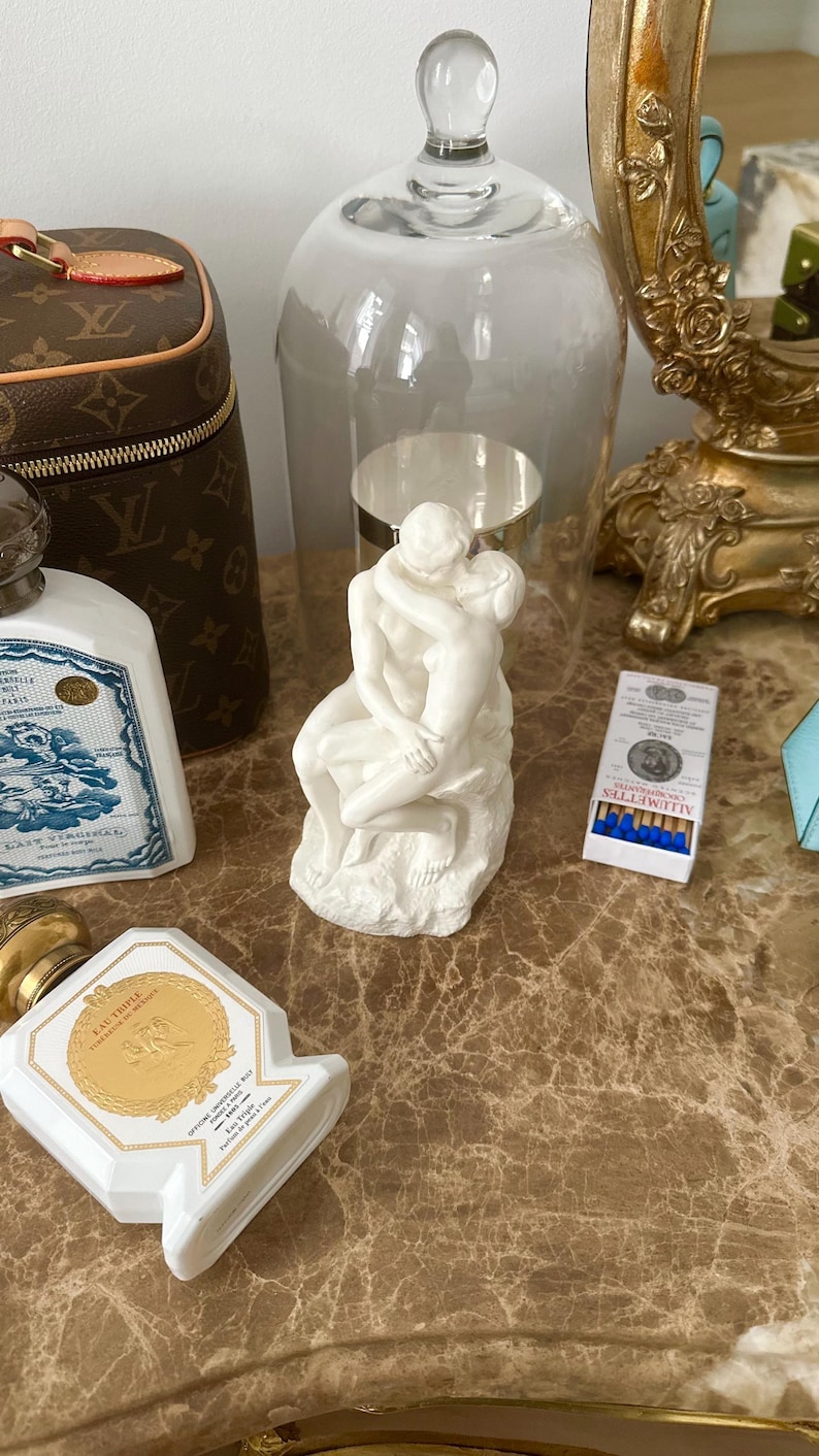 Peut inclure: Une sculpture en porcelaine blanche repr&eacute;sentant un couple en train de s'embrasser, expos&eacute;e sous un d&ocirc;me en verre sur une surface en marbre. La sculpture est entour&eacute;e d'autres objets d&eacute;coratifs, dont un flacon de parfum, une bo&icirc;te d'allumettes et un sac &agrave; main Louis Vuitton.