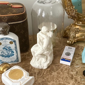 Peut inclure: Une sculpture en c&eacute;ramique blanche d'un couple s'embrassant, sous un cloche en verre, sur une surface en marbre avec un sac Louis Vuitton marron, un flacon de parfum et une bo&icirc;te d'allumettes.