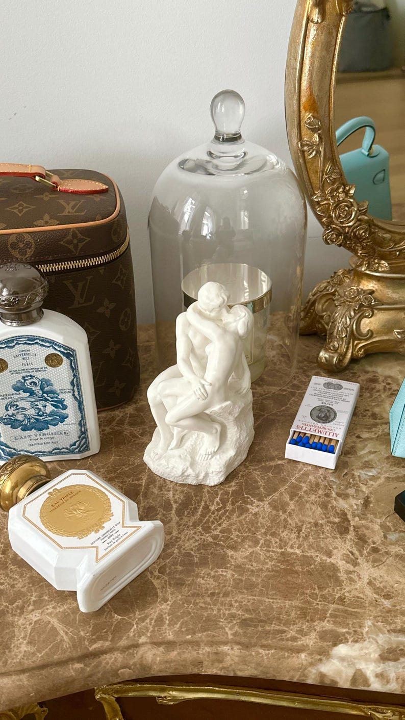 Peut inclure: Une sculpture en c&eacute;ramique blanche d'un couple s'embrassant, sous un cloche en verre, sur une surface en marbre avec un sac Louis Vuitton marron, un flacon de parfum et une bo&icirc;te d'allumettes.