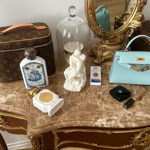 Peut inclure: Une table de toilette avec un plateau en marbre et des accents dor&eacute;s. La table pr&eacute;sente un petit sac &agrave; main bleu, une sculpture en porcelaine blanche, une cloche en verre transparent, deux flacons de parfum, un petit compact noir et un pinceau &agrave; maquillage dor&eacute;. La table est devant un miroir dor&eacute; avec un motif de ch&eacute;rubin.