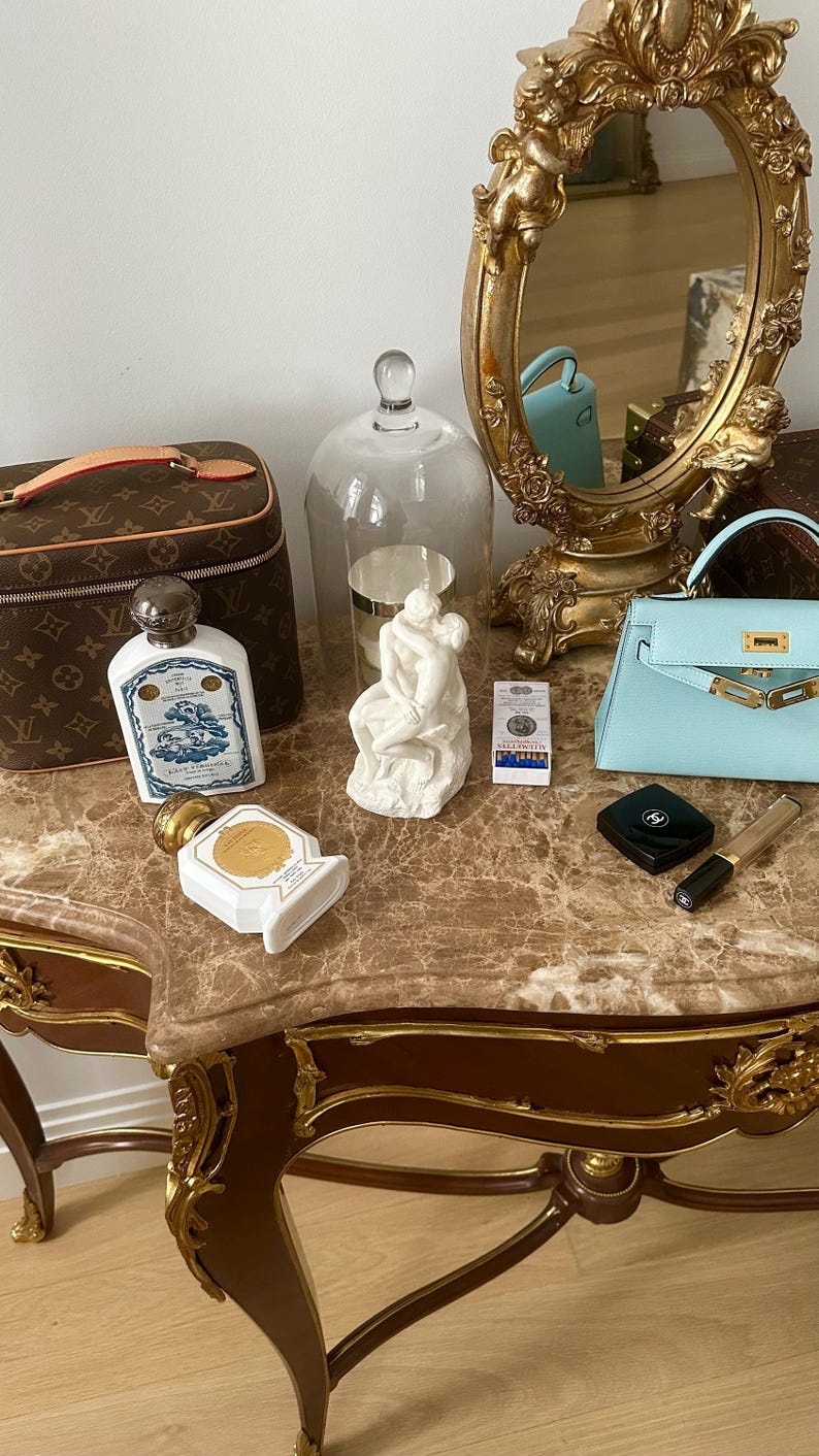 Peut inclure: Une table de toilette avec un plateau en marbre et des accents dor&eacute;s. La table pr&eacute;sente un petit sac &agrave; main bleu, une sculpture en porcelaine blanche, une cloche en verre transparent, deux flacons de parfum, un petit compact noir et un pinceau &agrave; maquillage dor&eacute;. La table est devant un miroir dor&eacute; avec un motif de ch&eacute;rubin.