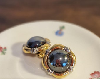 Könnte beinhalten: Ein Paar Ohrclips in Gold- und Silbertönen mit schwarzen runden Steinen. Die Ohrringe liegen auf einer weißen Untertasse mit Blumenmuster.
