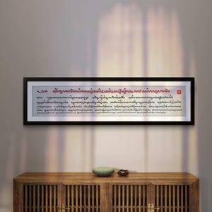 Puede incluir: Una obra de arte enmarcada con texto en escritura extranjera, expuesta sobre un mueble de madera. La obra de arte tiene un marco negro y un fondo blanco con texto rojo y negro. Un pequeño objeto de cerámica verde y un pequeño objeto marrón se encuentran sobre el mueble.
