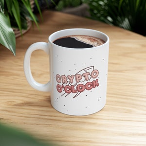 May include: White ceramic coffee mug with a black handle. The mug has a pink and white graphic of a rocket with the text "CRYPTO O'CLOCK".